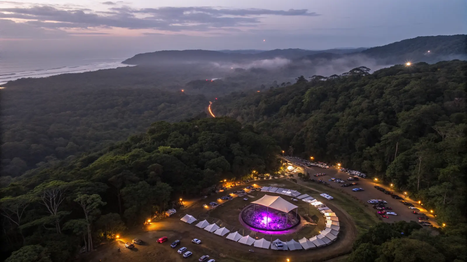 Vista aérea nocturna de un claro pequeño entre árboles con un escenario circular iluminado, carpas, carros y la costa a lo lejos